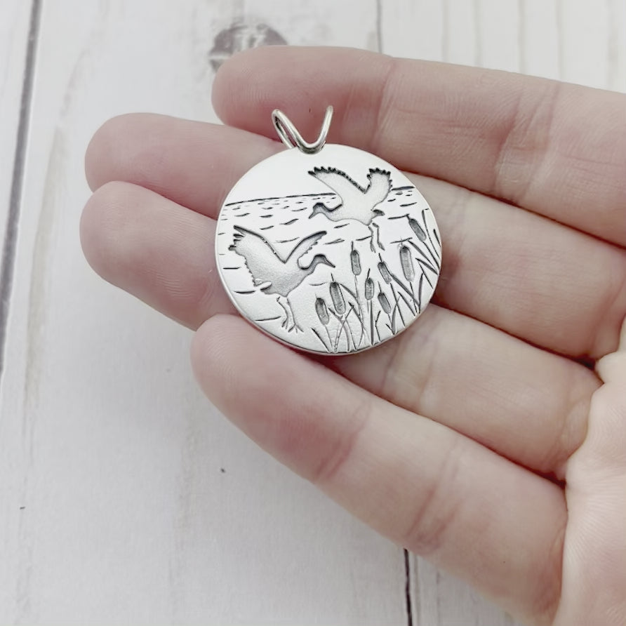 Round sterling silver pendant featuring two dancing cranes and cattail details. By Beth Millner Jewelry. 